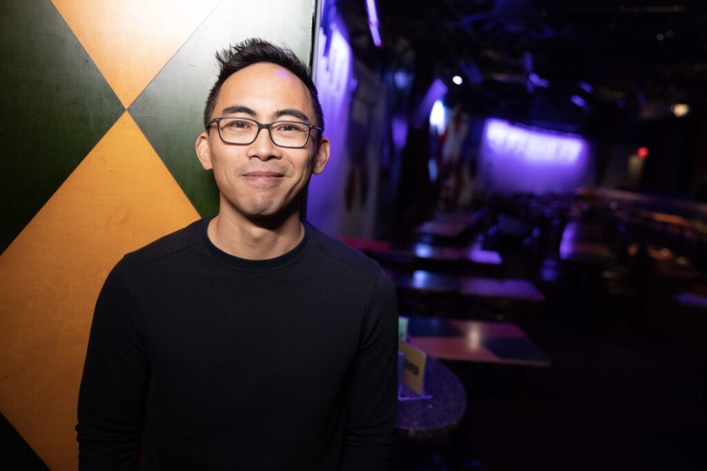 Photo of EJ Masicampo wearing a black long-sleeved shirt and glasses in front of a yellow and green checkered wall, with a stage behind him.