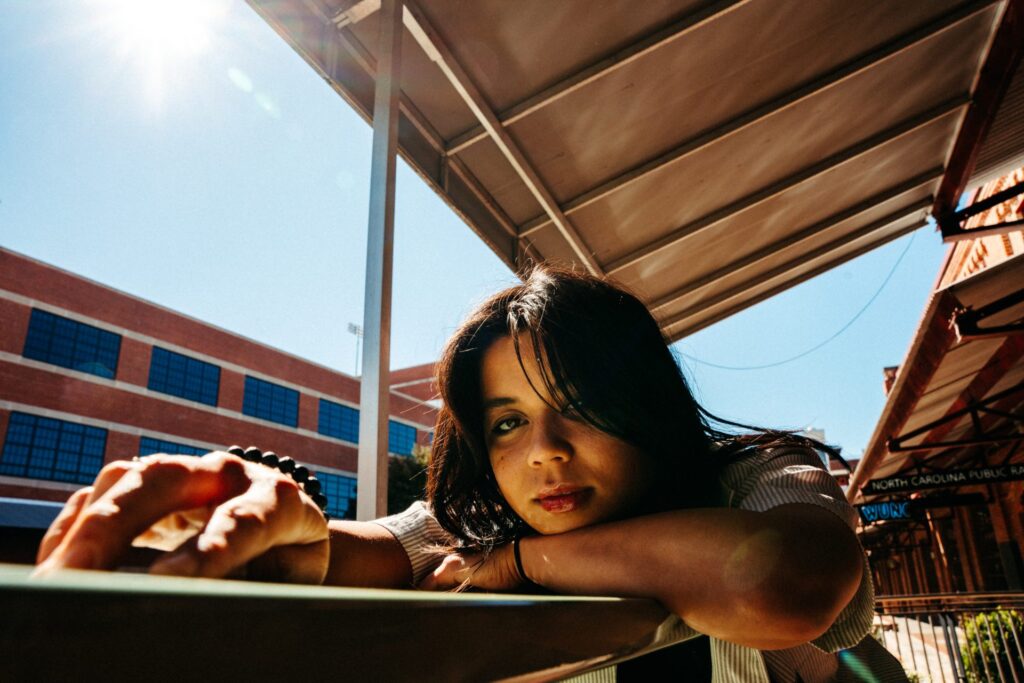 Naomi Poesel resting chin on arm on table. Her right hand is in the foreground, resting on the table. In the background, sunny clear sky and a patio-style roof.